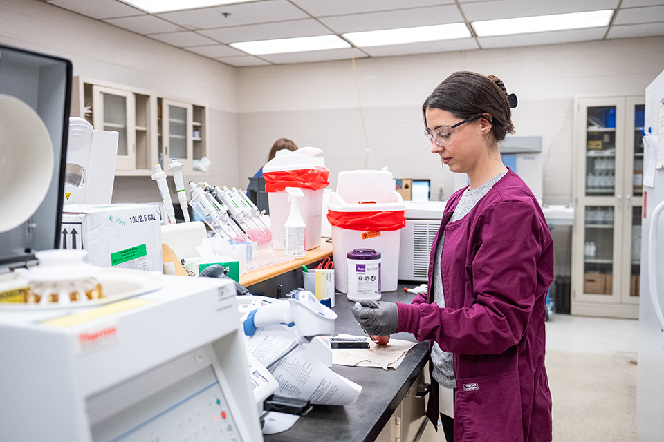 A woman in red scrubs prepares to test a sample in a diagnostic laboratory.