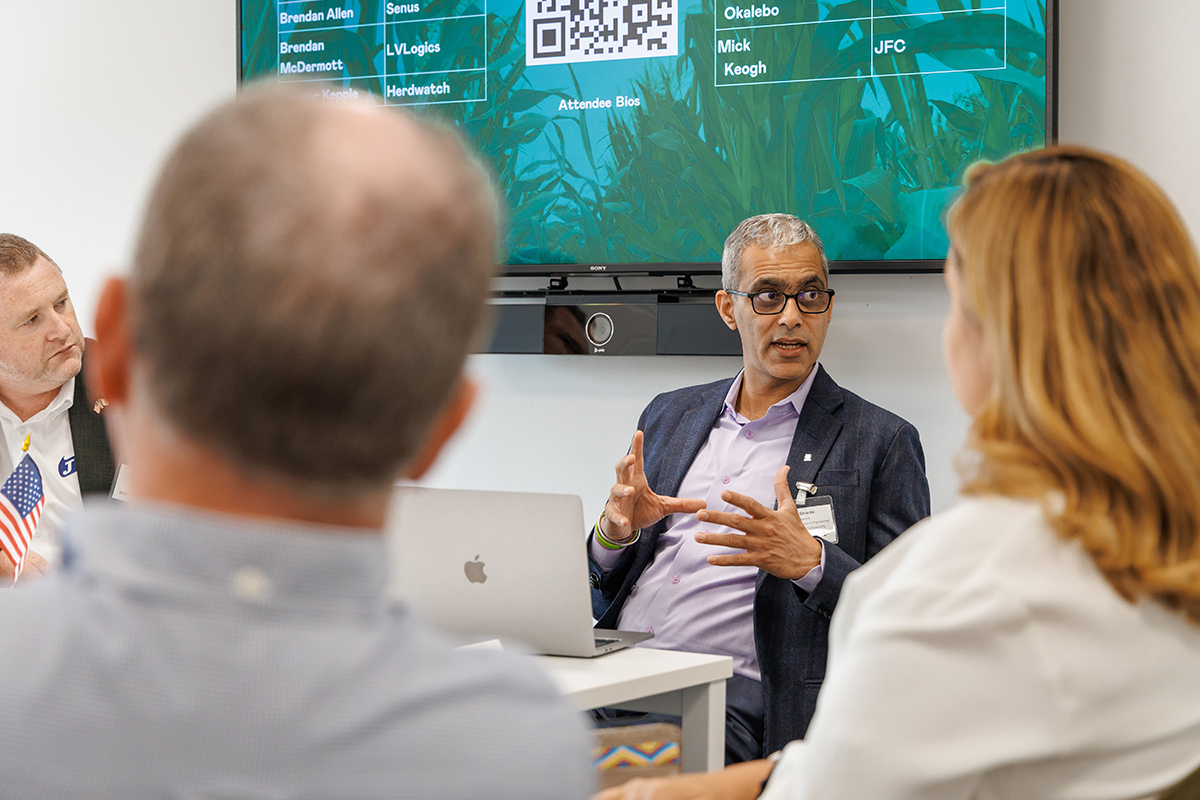 A man wearing glasses and a suit sits at a table and talks to people while gesticulating with his hands.