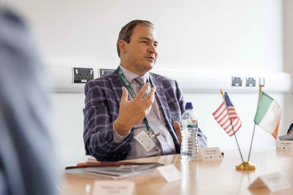 A man in a purple blazer sits at a table with American and Irish flags in front of him and gestures with his hand.