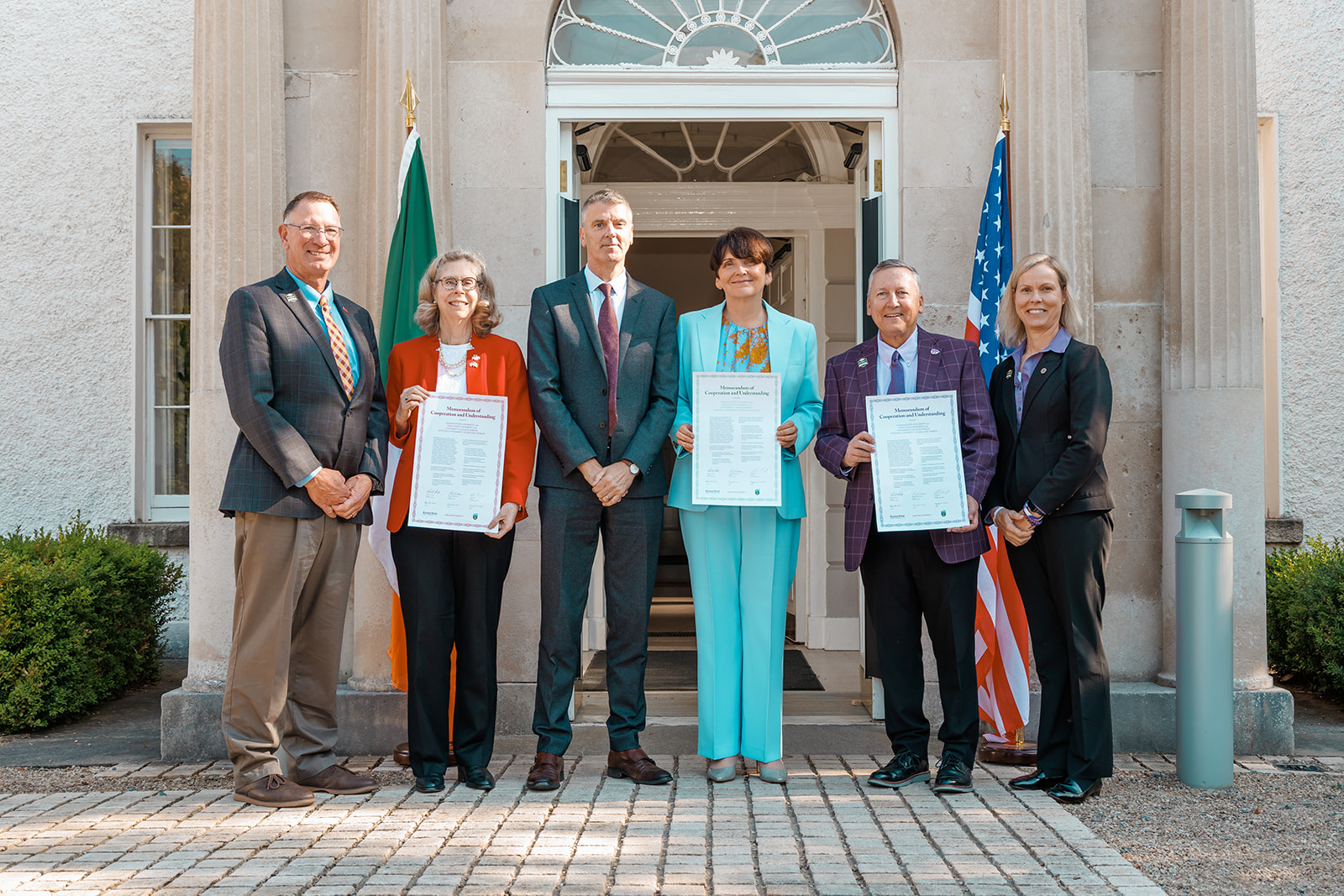 A group of six people in formal business attire stand in front of a building with the American flag and the Irish flag. Three people are holding up documents.