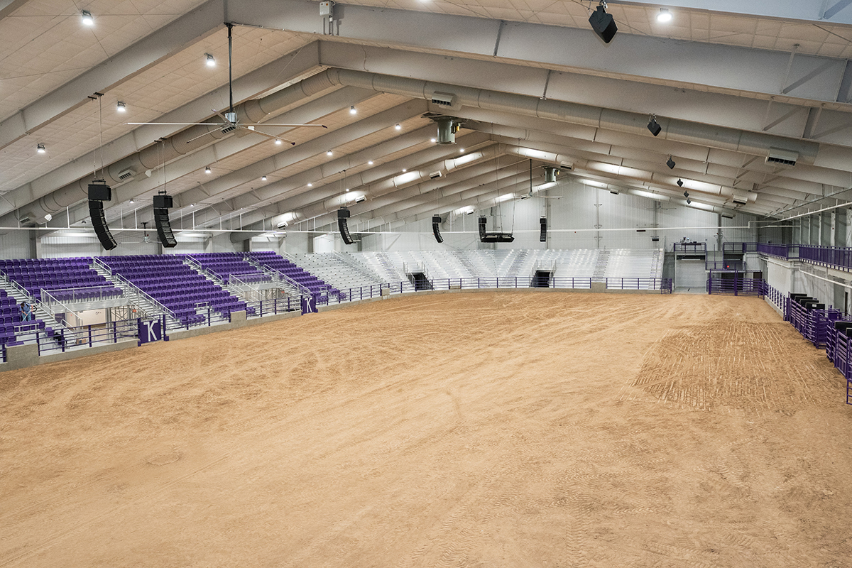 An indoor rodeo arena with sand-colored dirt and purple and white stadium seating surrounding.