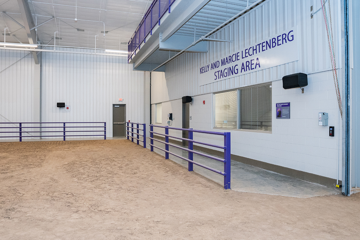 An indoor rodeo arena with dirt visible on the left; a staging area on the right of the photo has purple fencing and a gray wall with "Kelly and Marcie Lechtenberg Staging Area" in purple letters above a window.