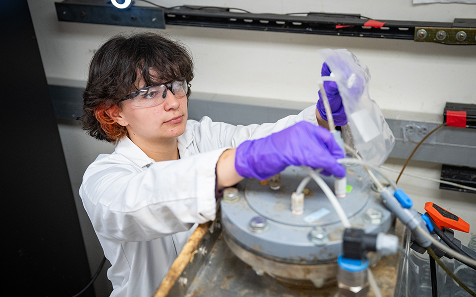 A college student wears a labcoat and purple gloves and works with a machine in a reseach lab.