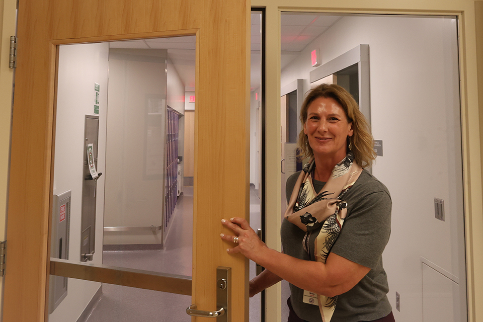 A college professor in a gray t-shirt holds open the door to a lab space.