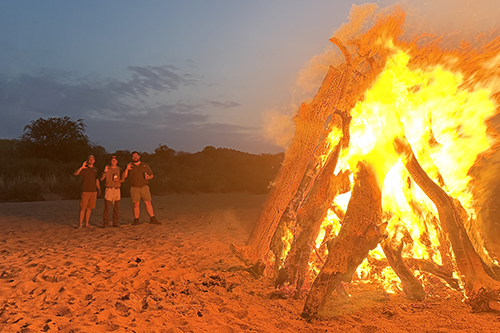Three men stand next to a large bonfire at night.