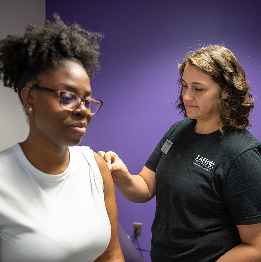 A close-up shows a pair of gloved hands injecting a flu vaccine into a person's arm.