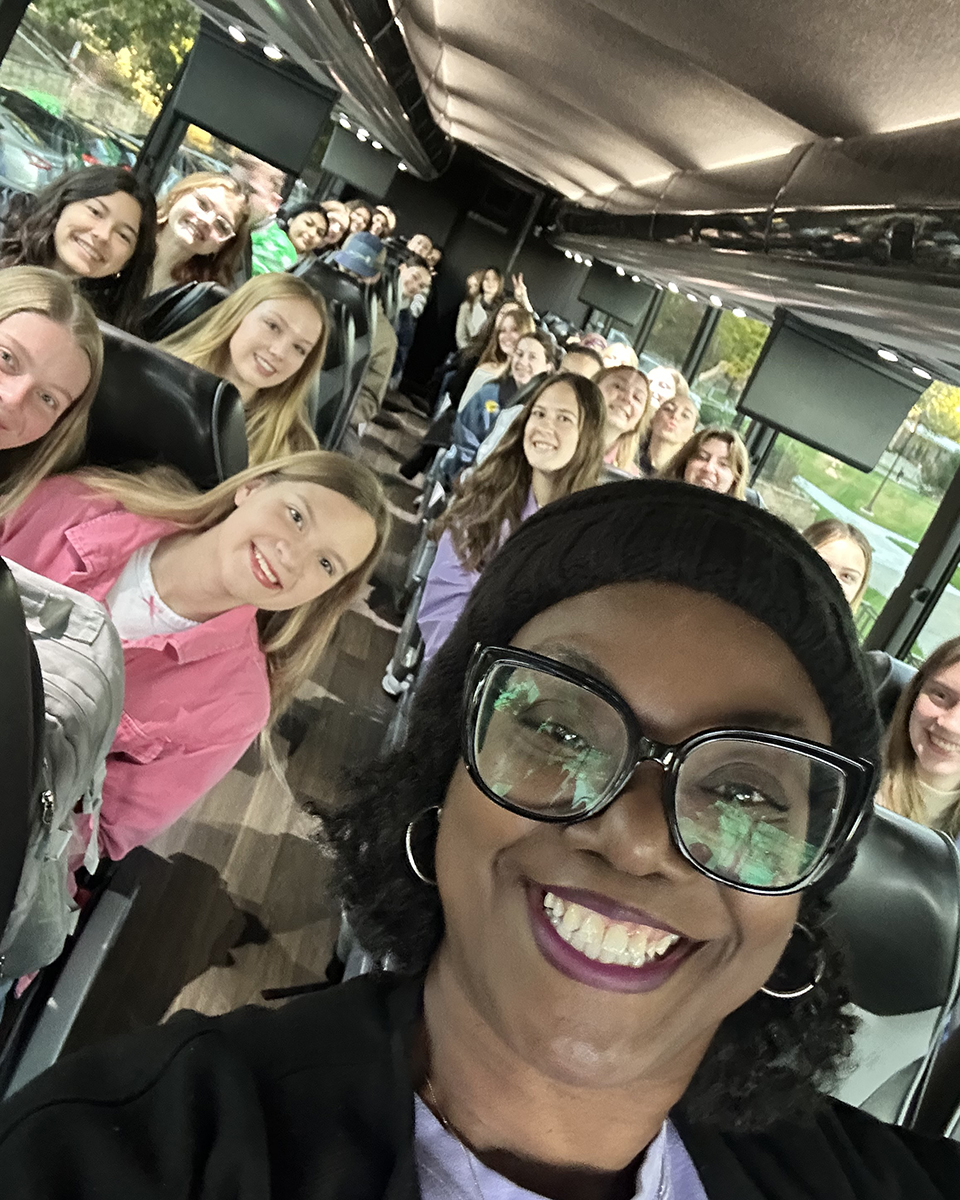A college advisor smiles and flashes a W-C hand symbol with a group of students on a bus.