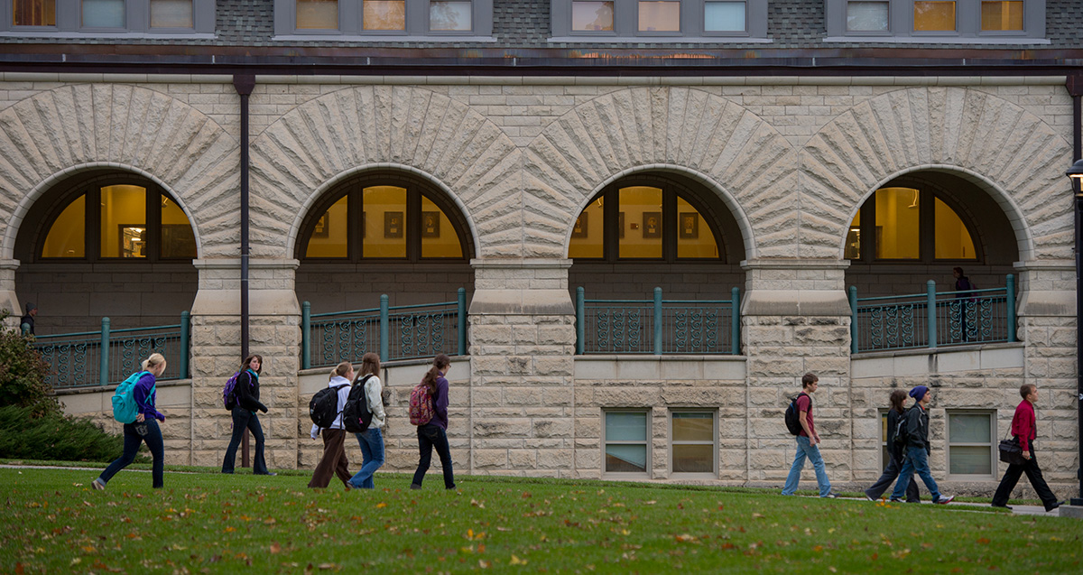 Six college students walk on a cement path through a lawn in front of a large limestone library with arches.