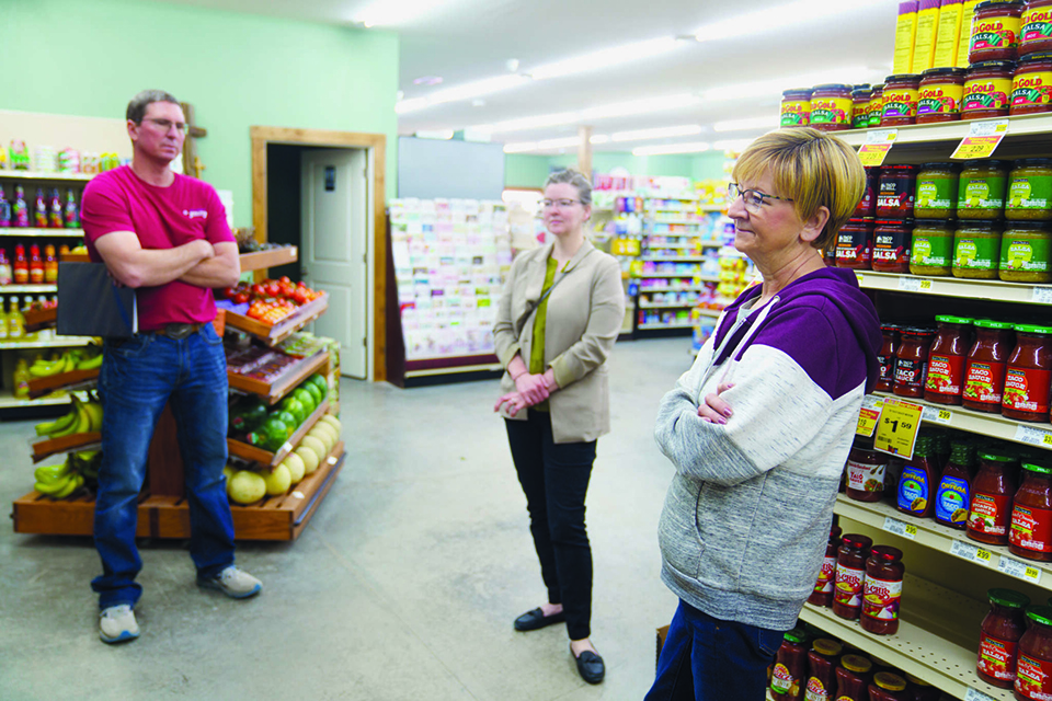 A group of community members stand and have a conversation beside a shelf of groceries in a store.