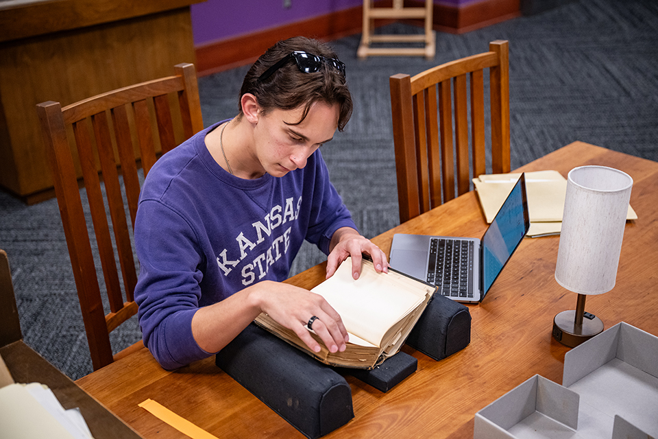 A student wearing sunglasses on the top of their head and a purple "KANSAS STATE UNIVERSITY" shirt sits at a wooden library table and carefuly reads through an old book. A laptop sits next to the book.
