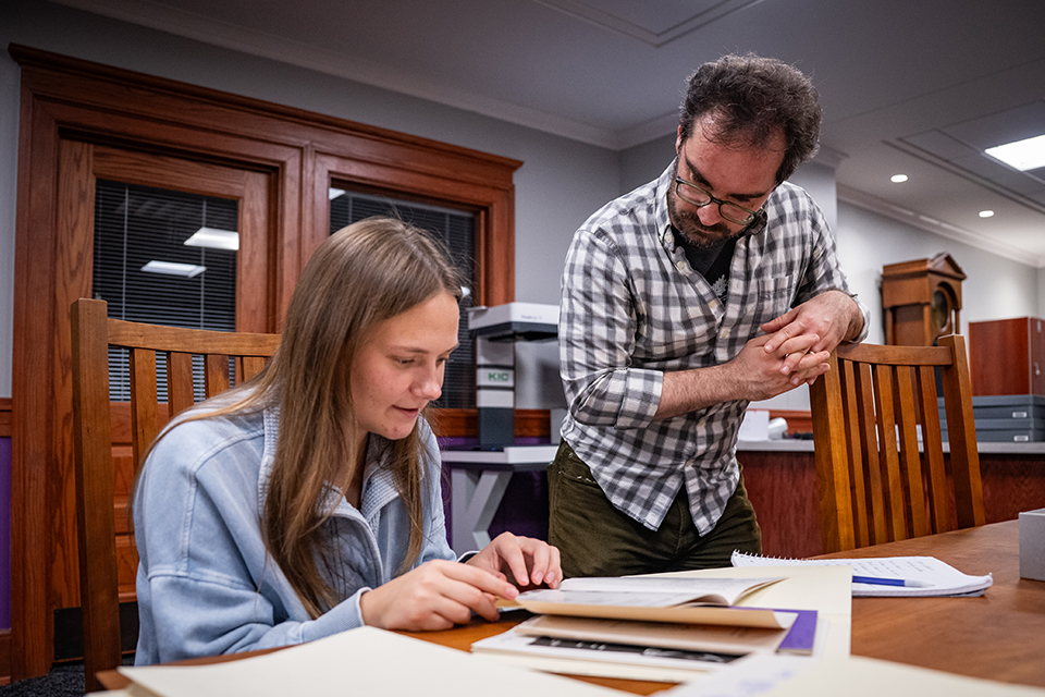 A college professor looks over a student's shoulder as she sits at a library table and looks through an antique book.