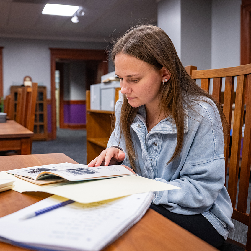 A female college student sits and combs through an old collection of Greek life photos at a wooden library table.