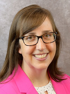 A woman with brown hair and glasses wearing a pink blazer and white polka dot shirt smiles for a portrait photo.