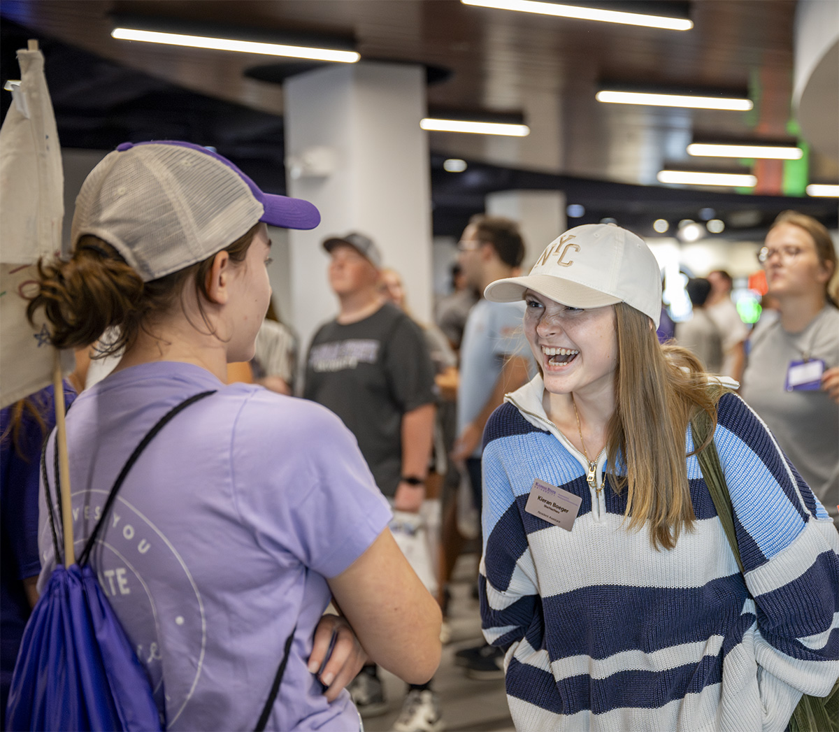 A college student in a blue striped sweater and a white and tan hat smiles toward another student in a purple t-shirt and a purple and gray mesh hat.