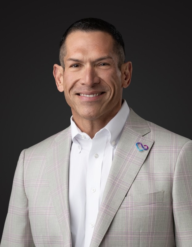 Daniel Akina smiles in a white button-up and tan plaid suit for a professional portrait.