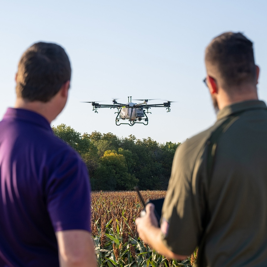 Two men look at and operate an agricultural spraying drone that flies over a field on a sunny, clear day.