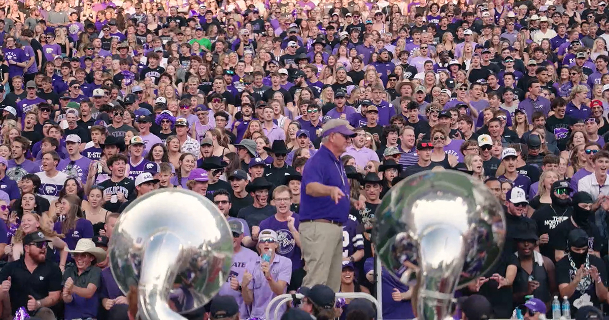 A marching band director stands between a section of tubas and a student section at a football game.