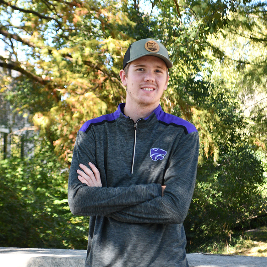 A college student wears a baseball hat and sweatshirt and poses for a portrait outside against a blurry green foliage background. 