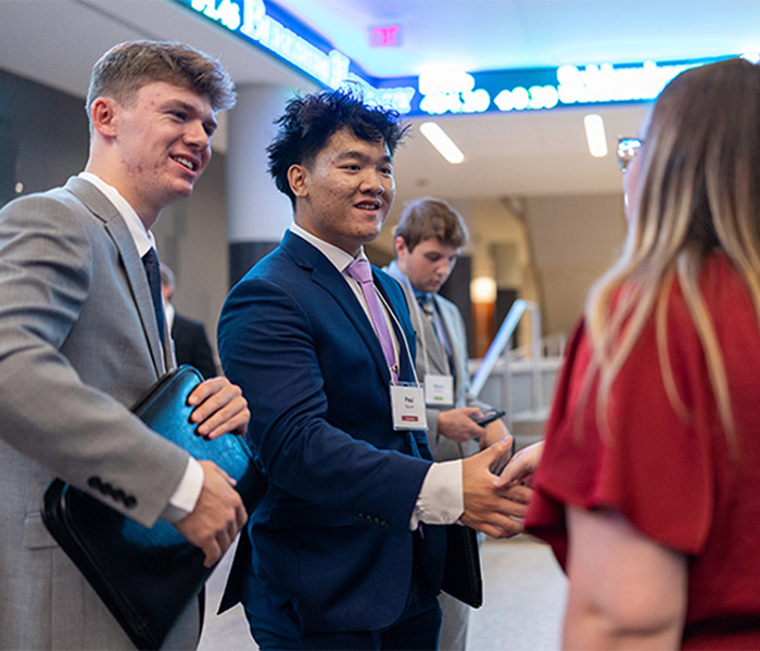 Two students in business attire greet and shake hands with an attendee at a professional networking event inside a campus building.