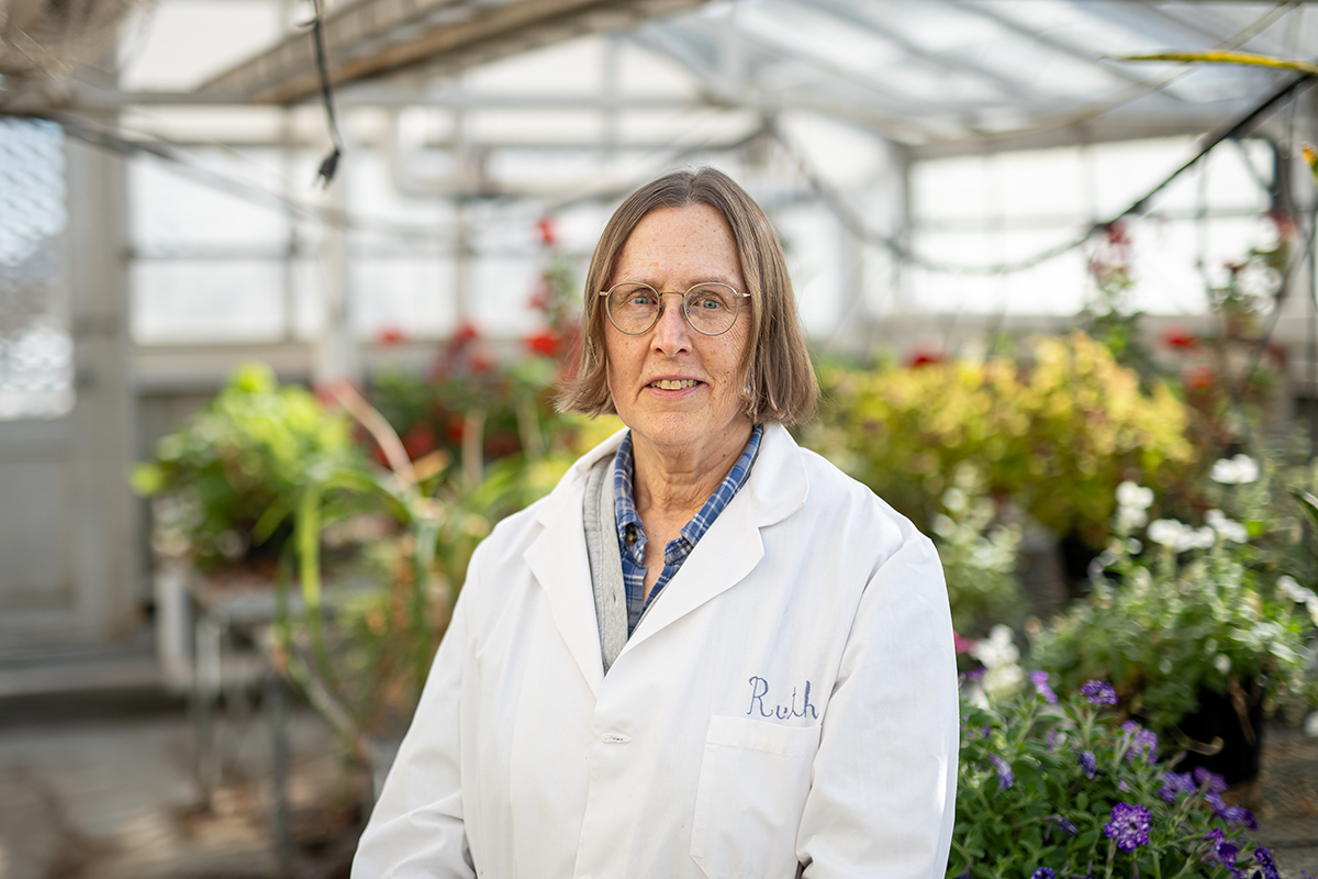 Ruth Welti wears a white lab coat and smiles for a professional portrait in a brightly lit greenhouse filled with different flowering plants. 