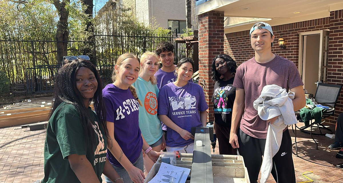 Madeleine Pike stands to the left wearing a purple K-State shirt, and Roberto Maldonado Flores stands to the right, also wearing a purple shirt, with other students in Dallas, Texas.