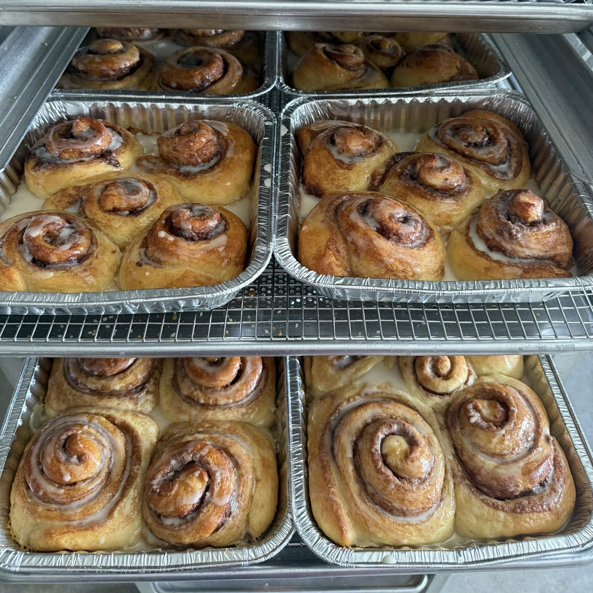 Several trays of baked, glazed cinnamon rolls sit on cooling racks.