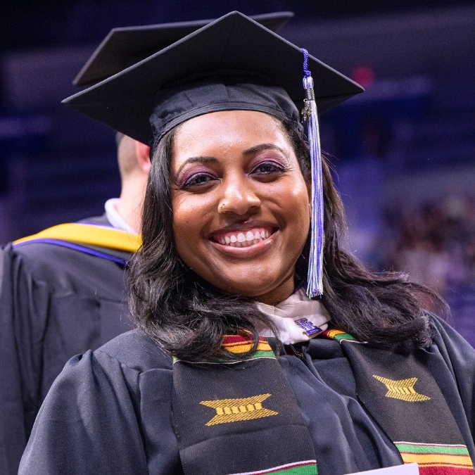A Black woman is pictured wearing a graduation cap and gown with a stole with red, yellow and green markings.