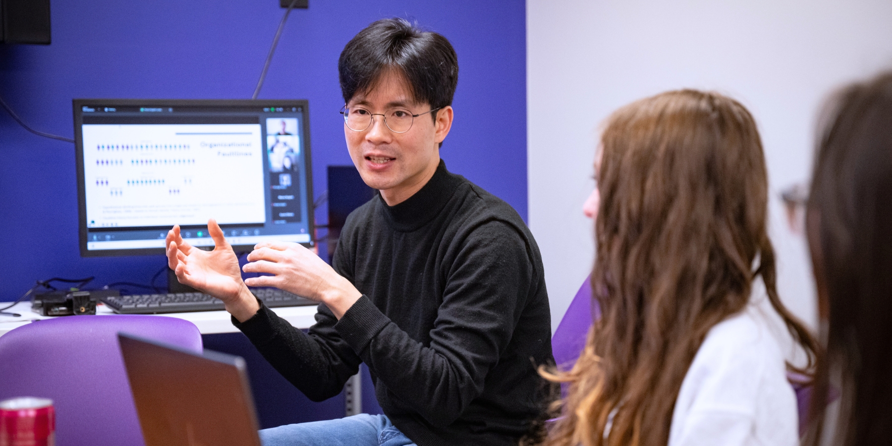 Jin Lee teaches students with his hands raised in a natural speaking position. He is sitting in a purple chair and wearing a black turtleneck. In the background, there is a purple wall and a computer screen.