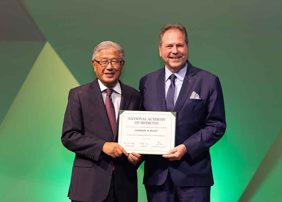 Two men in suits pose for a photo and stand next to each, both holding the same certificate.
