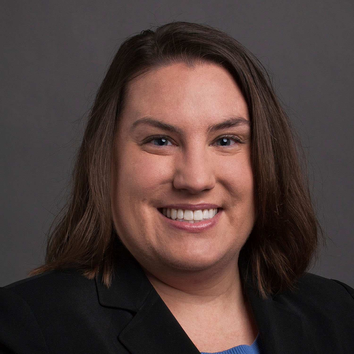 A close-up portrait photo shows a woman's shoulders and face. She is wearing a back blazer and has shoulder-length brown hair.