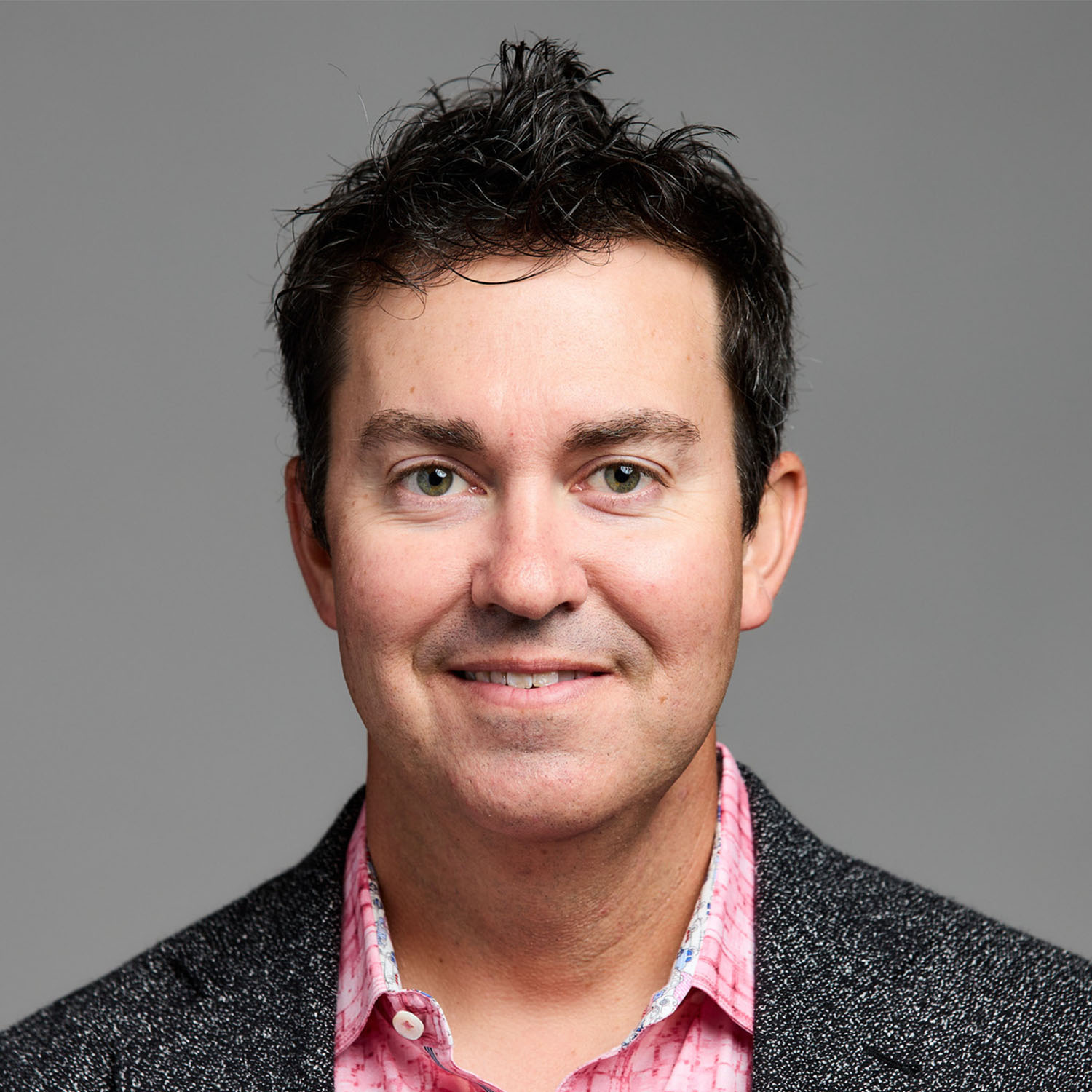 A man with black hair smiles for a portrait photo, wearing a black blazer with a pink collared shirt