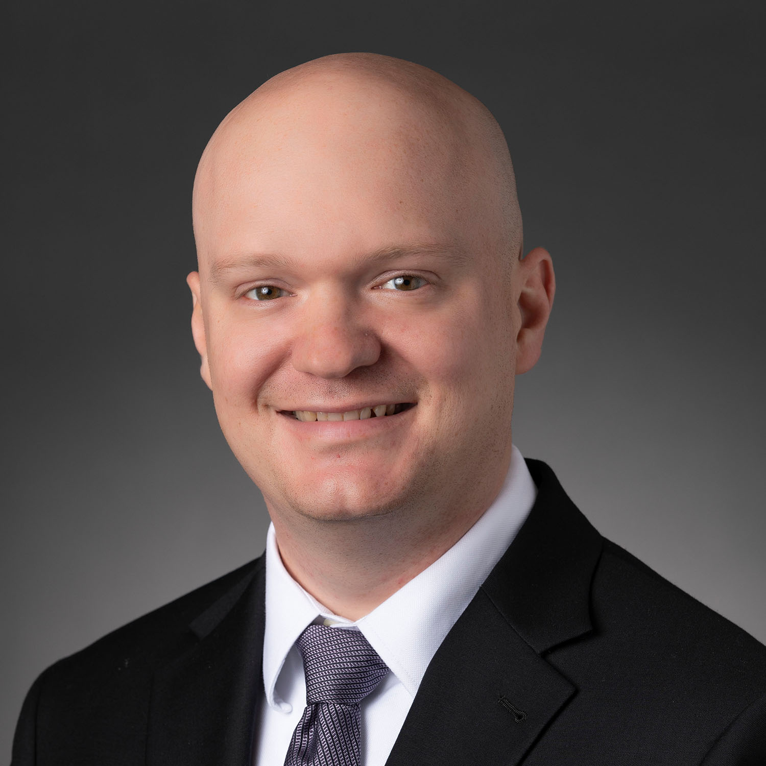 A bald man wearing a black blazer over a white collared shirt and a silver tie smiles against a gray background.