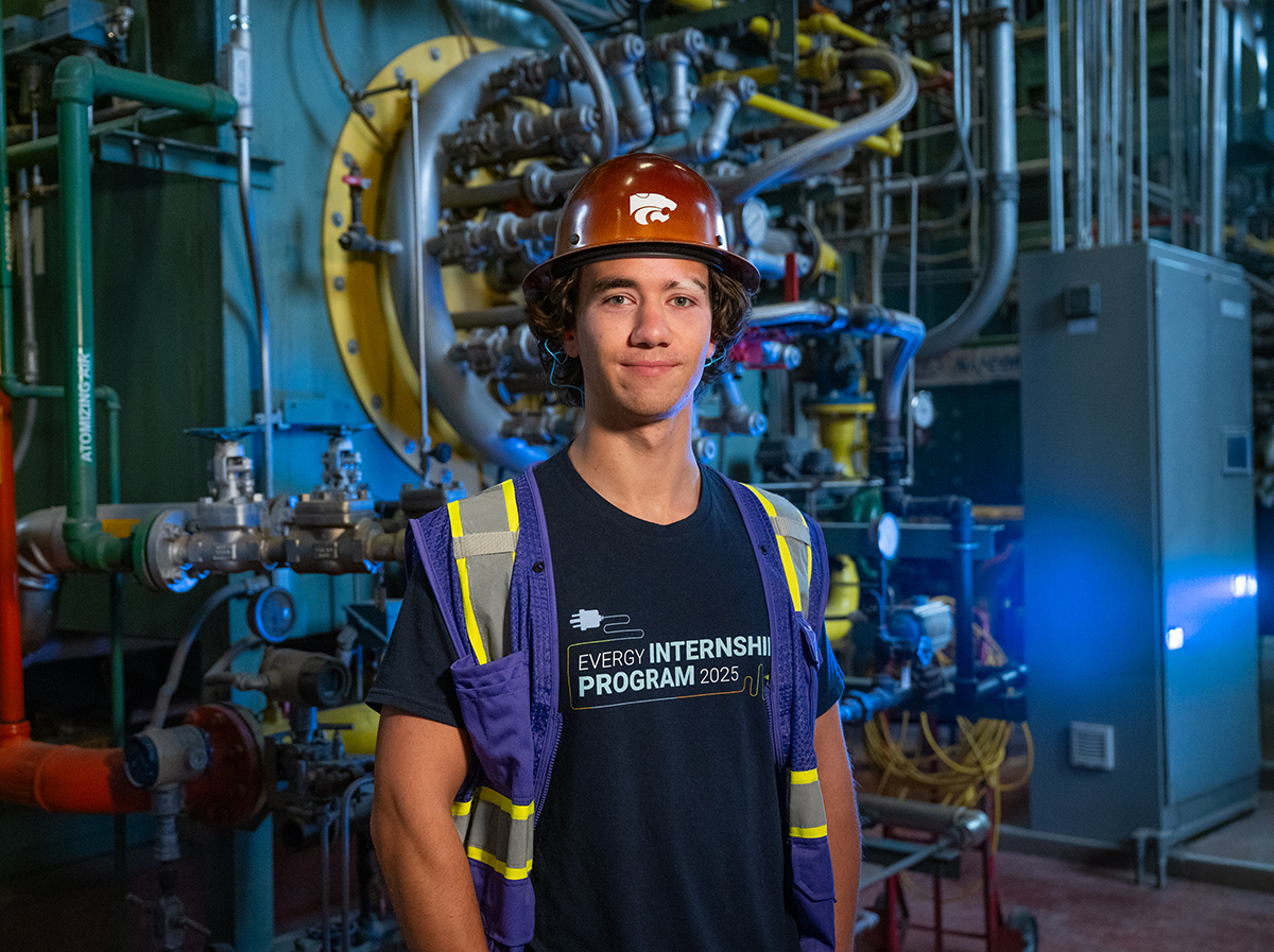 A young man with an orange hard hat with a powercat on it and a purple safety vest stands in front of an industrial background with lots of pipes and wiring.