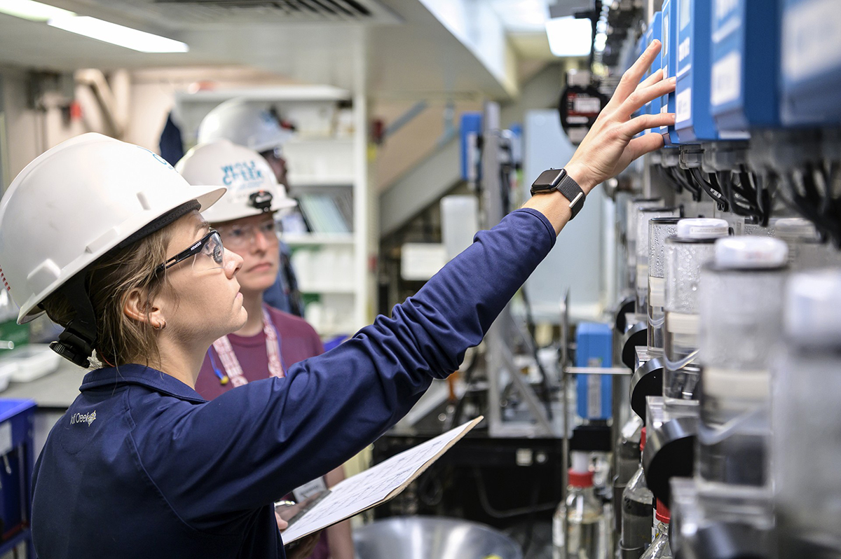 A young woman with brown hair in a white hard hat wearing a dark blue jacket reaches upward toward a blue box. Another intern in a white hard hat stands behind her observing.