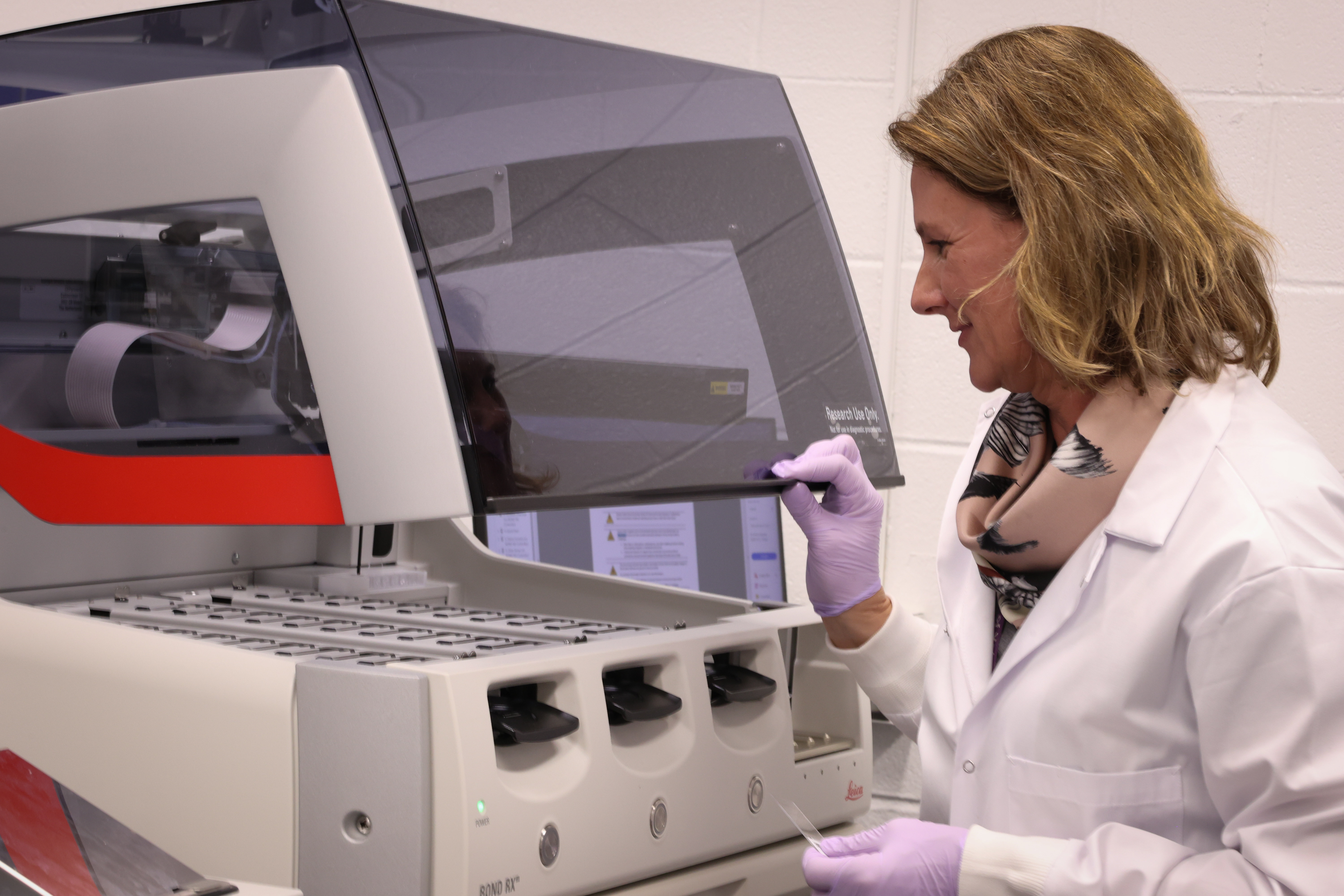 A woman wearing a white labcoat and lavender gloves touches a machine with a dark hood and several switches.