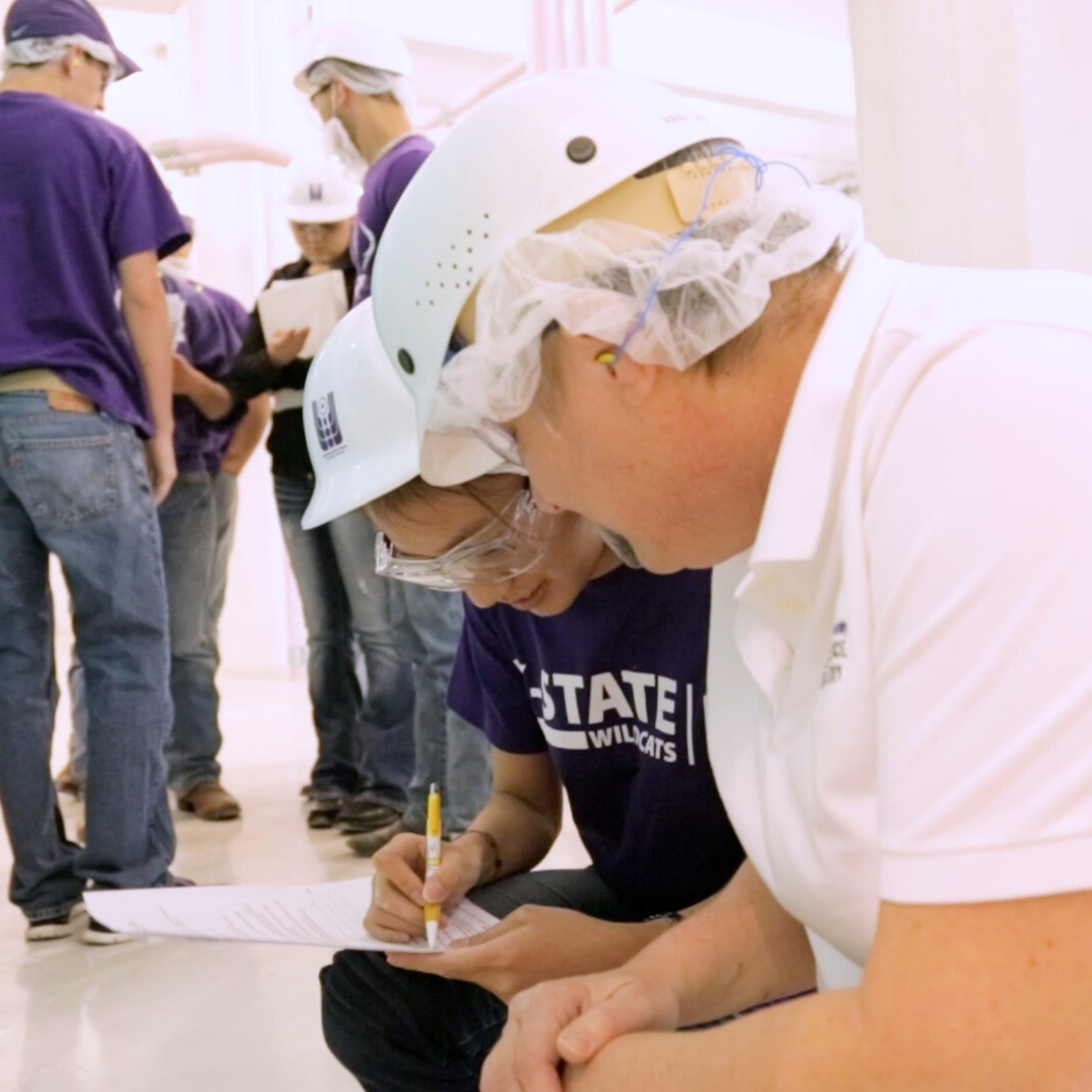 A female student and male teacher, both wearing white hardhats and goggles, crouch town to make note of research in the facility. The student, on the left, holds a yellow pen and writes down notes on a sheet of paper.