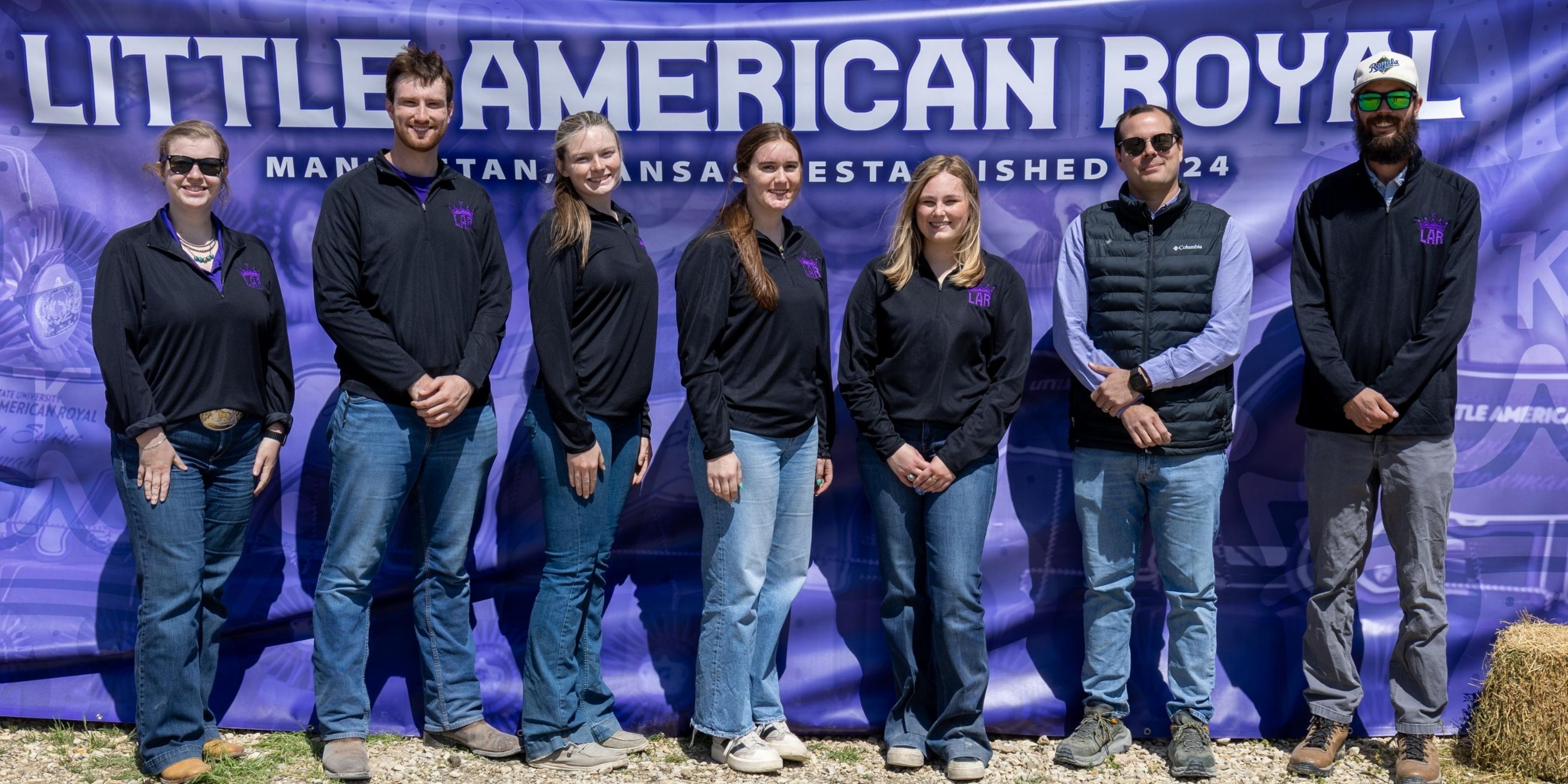 A group of students, including Carson Corwin, stand in a line in front of a purple banner reading 'Little Americal Royal." They all wear a black zip-up jacket and blue jeans.
