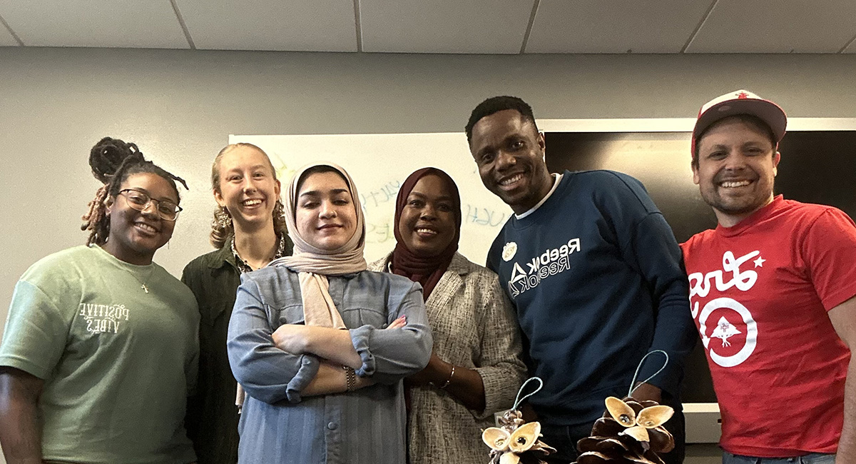 A group of college students and a staff member pose around a conference table in a campus meeting room.