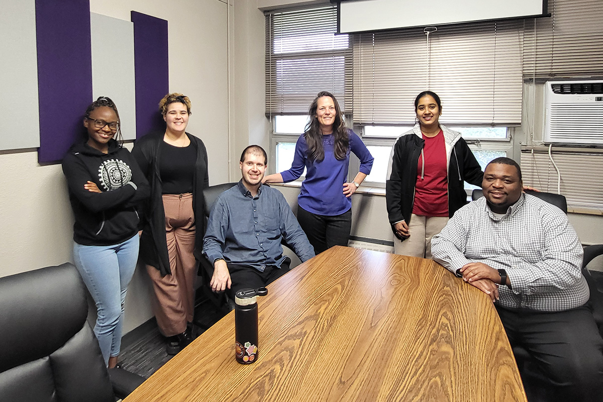 A group of college students and a staff member pose around a conference table in a campus meeting room.
