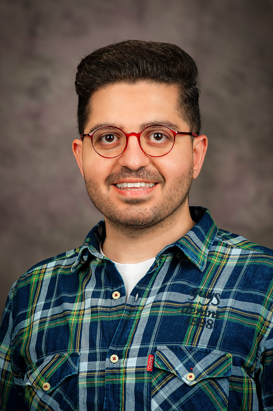 A graduate student with short black hair wearing a blue and green plaid shirt and red, round glasses smiles for a portrait photo against a gray backdrop.