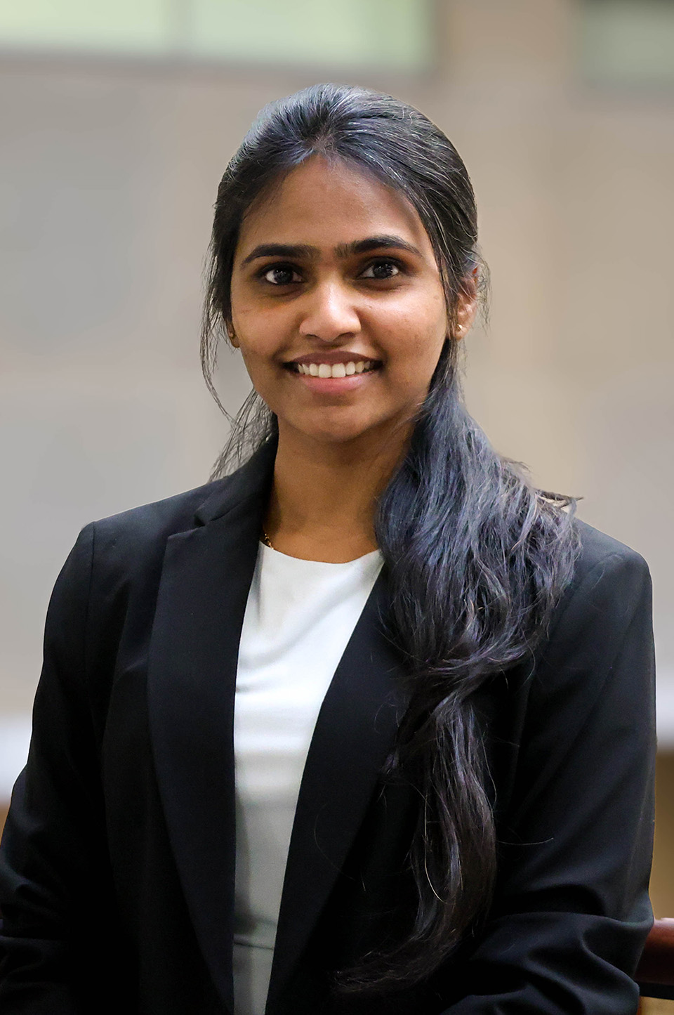 A graduate student with long dark hair in a ponytail wears a black blazer over a white shirt.
