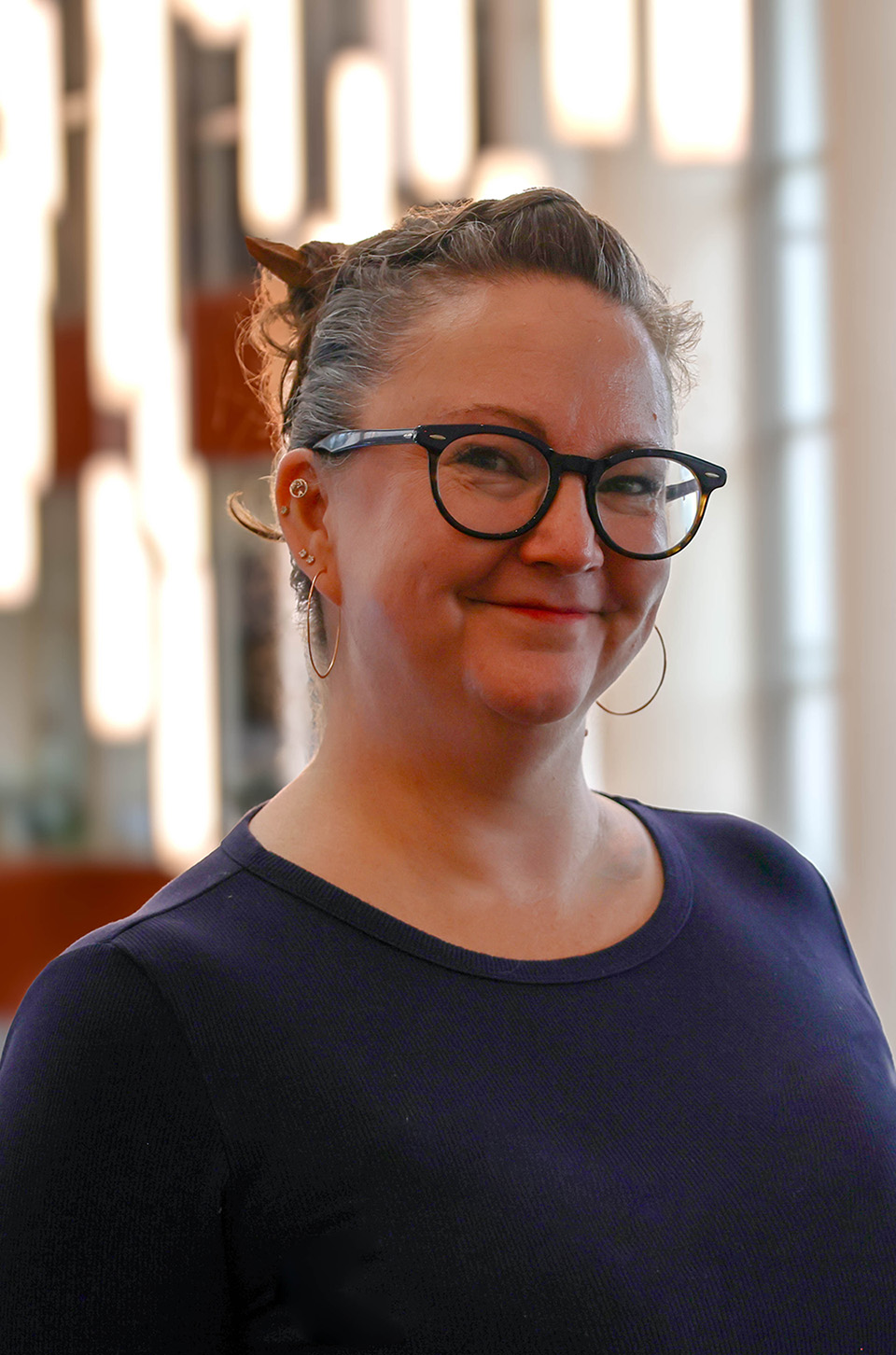 A graduate student with hair pulled back into a bun wears a dark purple shirt and glasses and smiles against a background of hanging lights.