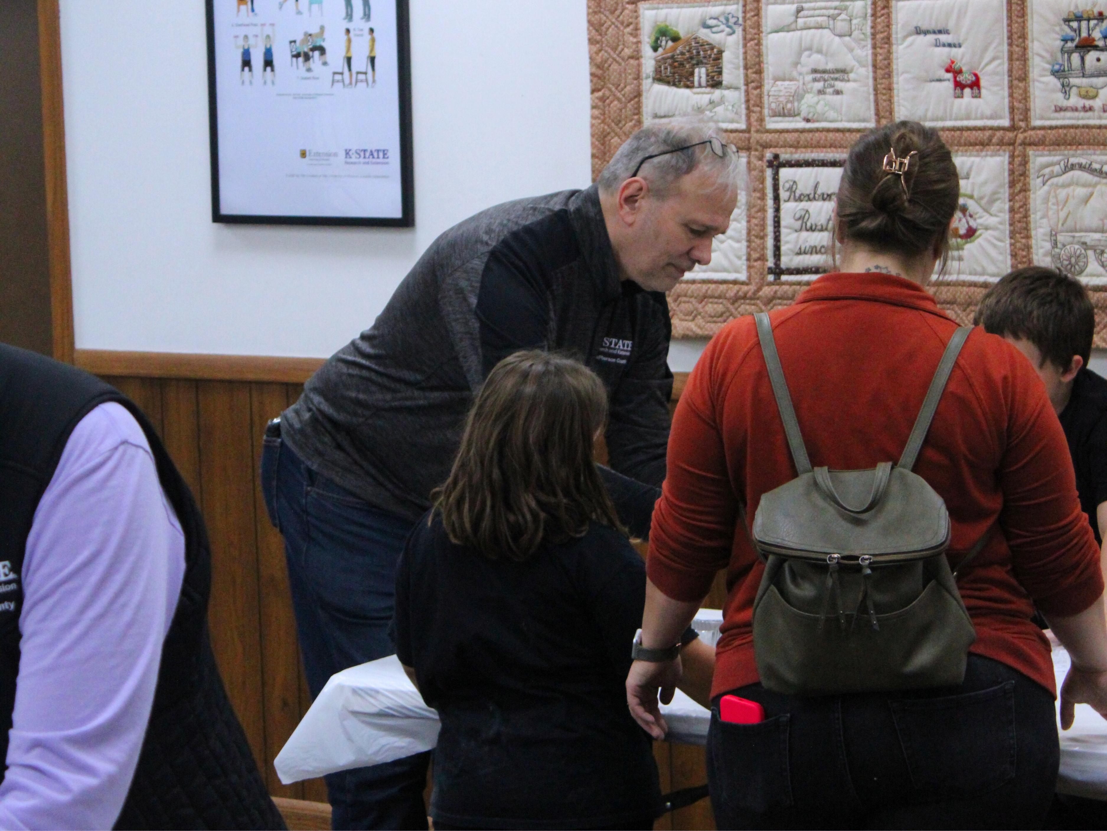 Chuck Laughlin, wearing a black and grey quarter-zip sweatshirt, leaves over a table to explain an activity to participants. In the foreground, there is a woman wearing a red shirt and grey drawstring bag and a girl wearing a black t-shirt.
