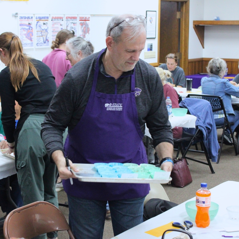 Chuck Laughlin, wearing a purple K-State Extension apron, offers supplies for a workshop on a baking sheet to participants. 