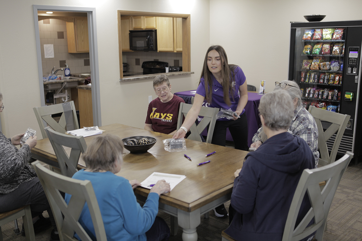 A young woman hands out pieces of paper at a table where several older women from a retirement community are sitting.