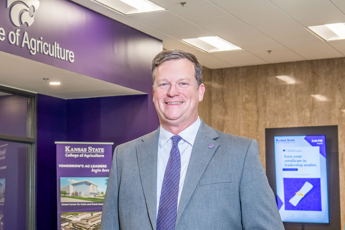 A male college dean in a gray suit and purple tie poses for a portrait in a college hallway outside of an office that has a purple powercat logo and the name 