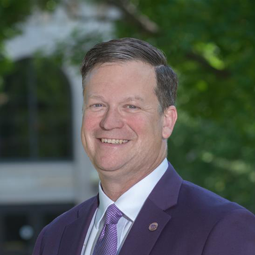 A male college dean in a purple suit and purple tie poses for a portrait. A purple background with the seal of Kansas State University is behind him.