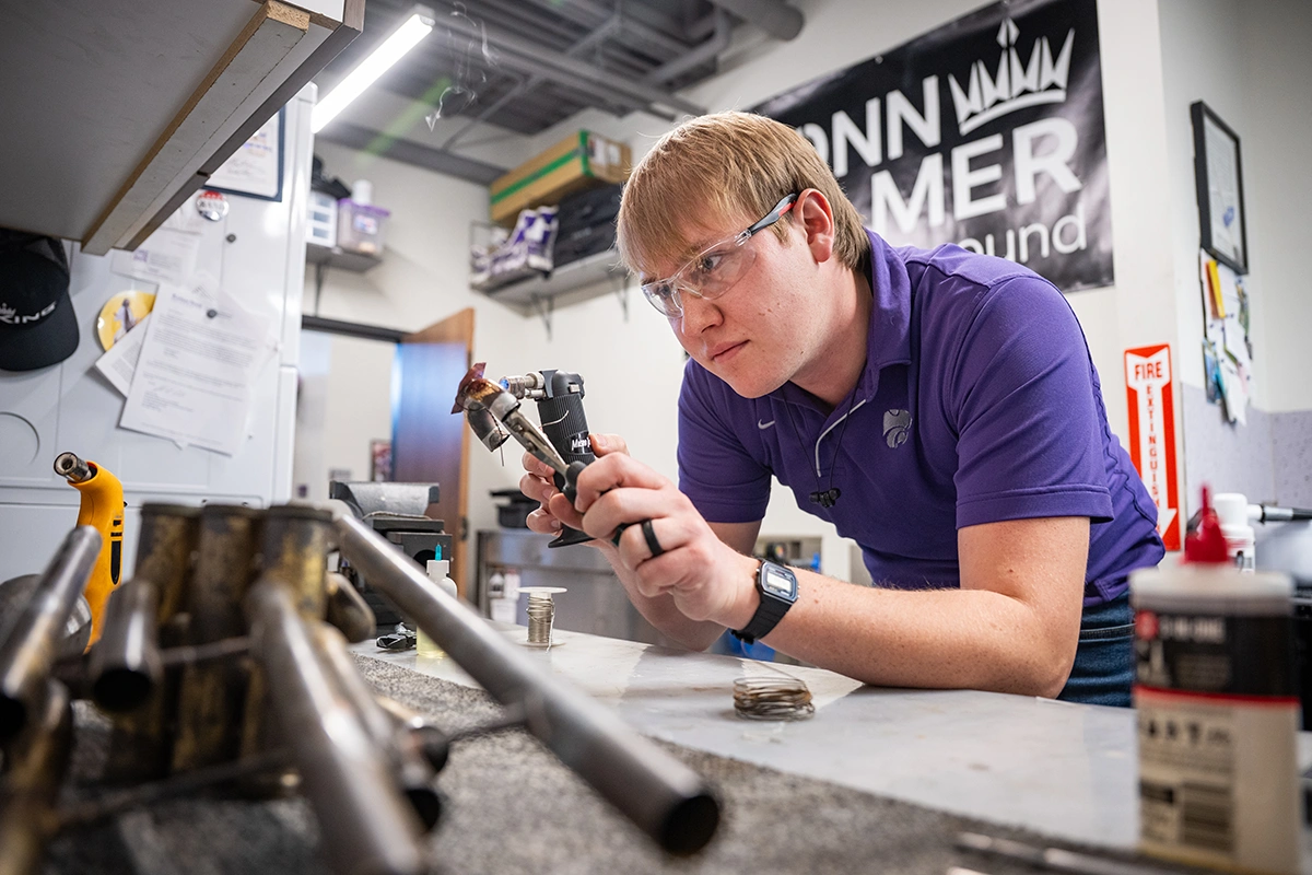 A male college student in a purple polo works with pieces of metal at a music instrument repair workbench.