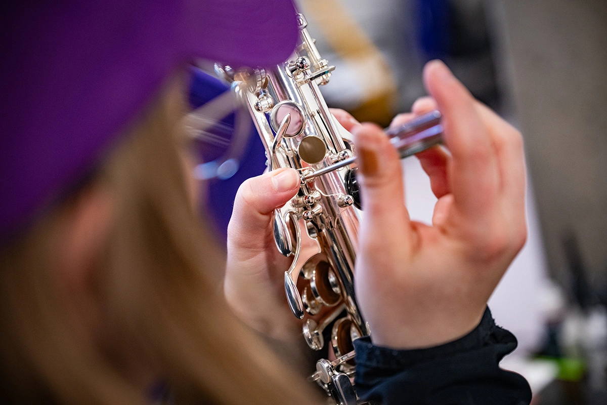 From the perspective over a female college student's shoulder, a pair of hands carefully remove the keys from a shiny metal saxophone.