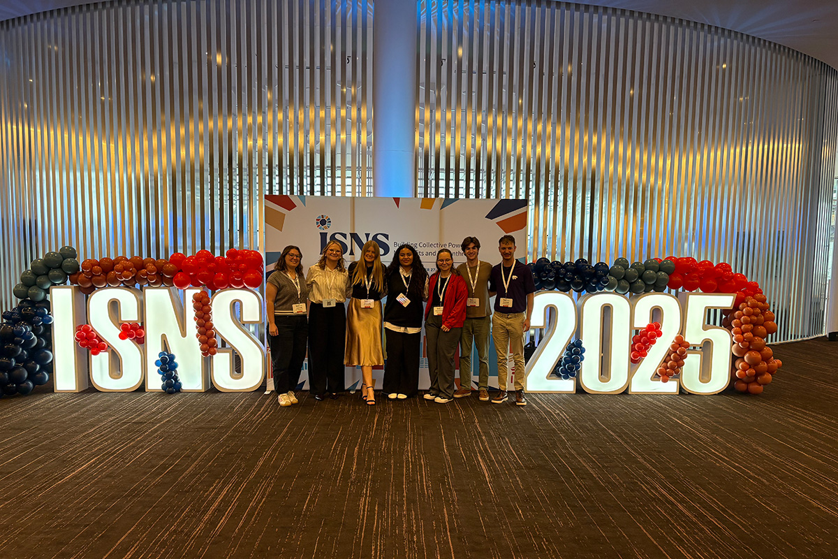 Seven college students stand in front of a photo backdrop with 